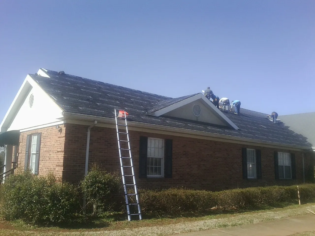 Crew tearing shingles off a brick ranch roof with a ladder set — Metal Roof Repair work by our crew in Sarahsville