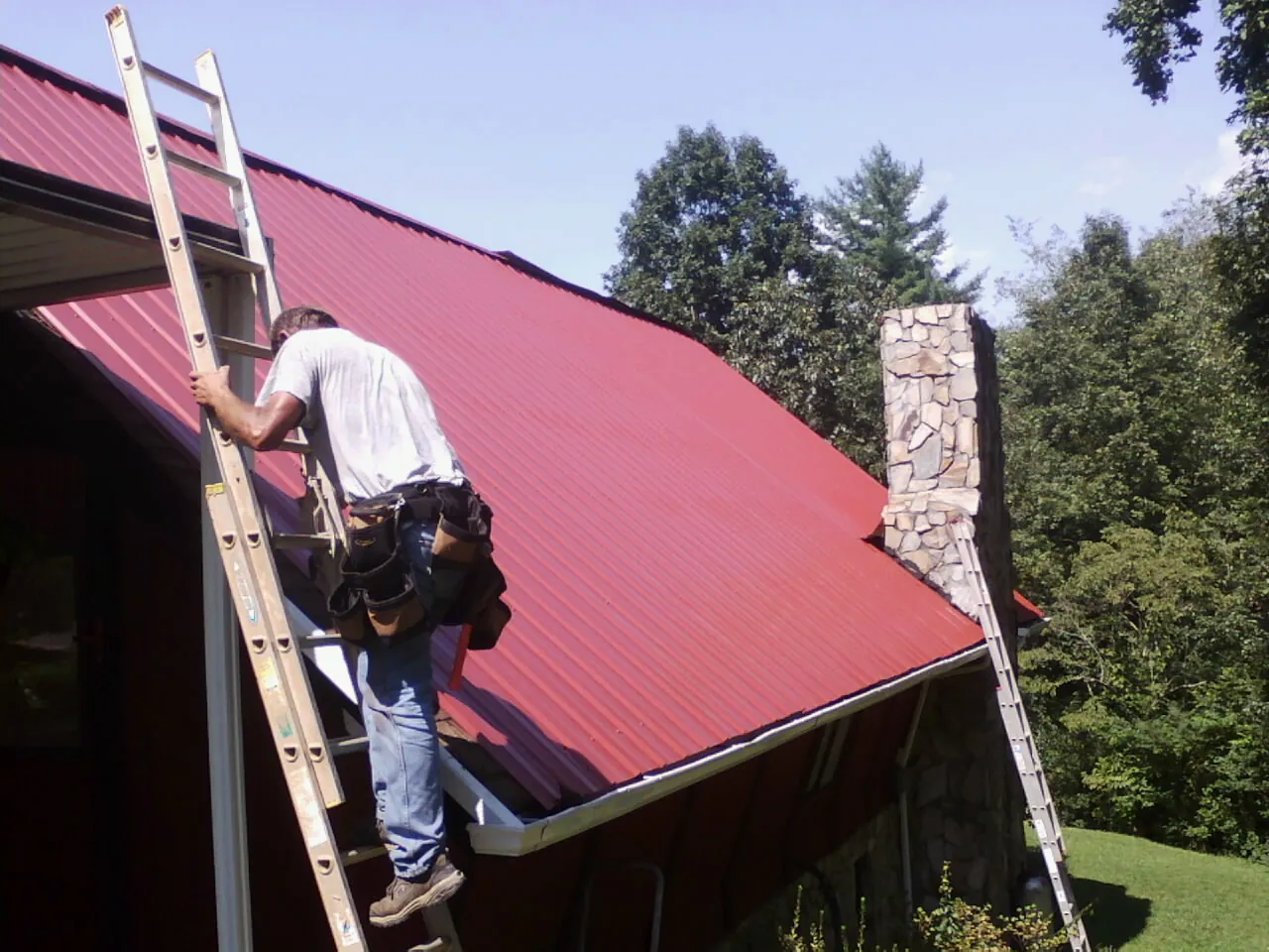 Roofer on a ladder beside a red metal roof and stone chimney — Soffit & Fascia Repair by Roofing Brothers in Sarahsville